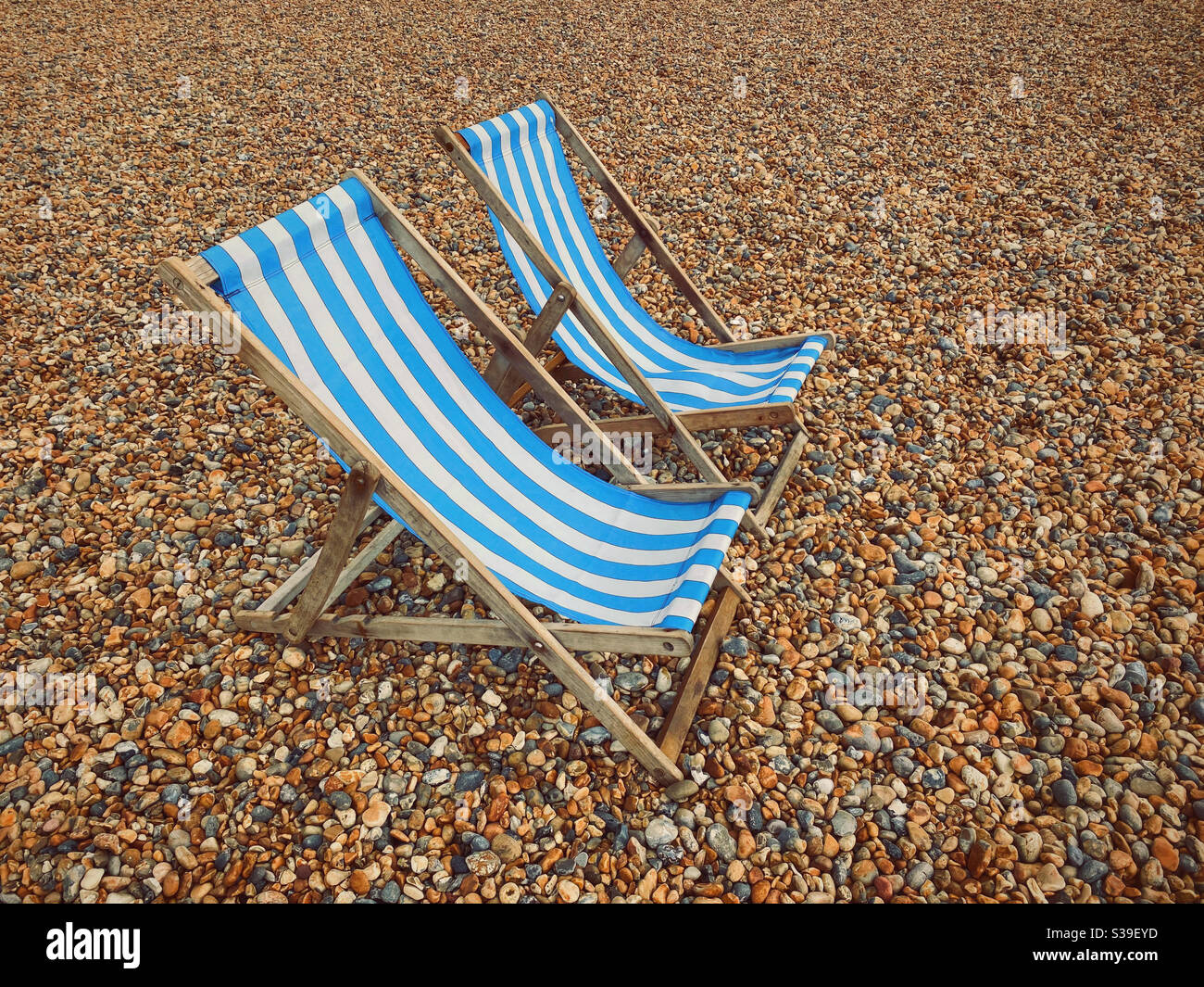 Due sedie a sdraio in legno a strisce blu e bianche, tutte da sole e circondate da una spiaggia di ghiaia. Dove sono tutte le persone? Foto ©️ COLIN HOSKINS. Foto Stock