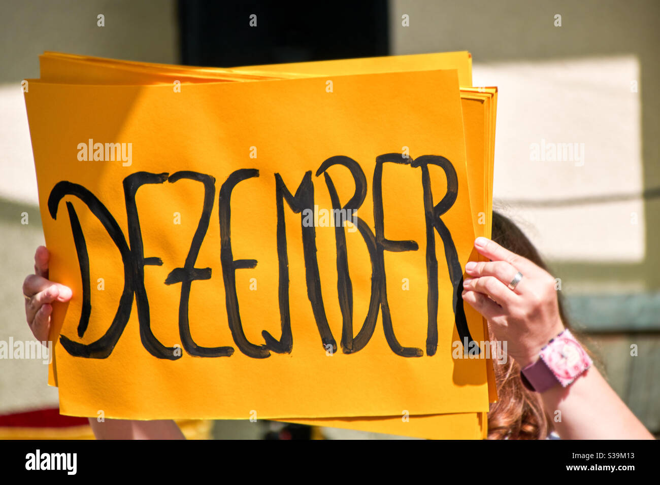 Lettere dipinte di nero formano la parola dicembre in tedesco su un cartone giallo come sfondo. Il cartello è tenuto in aria e illuminato dal sole Foto Stock