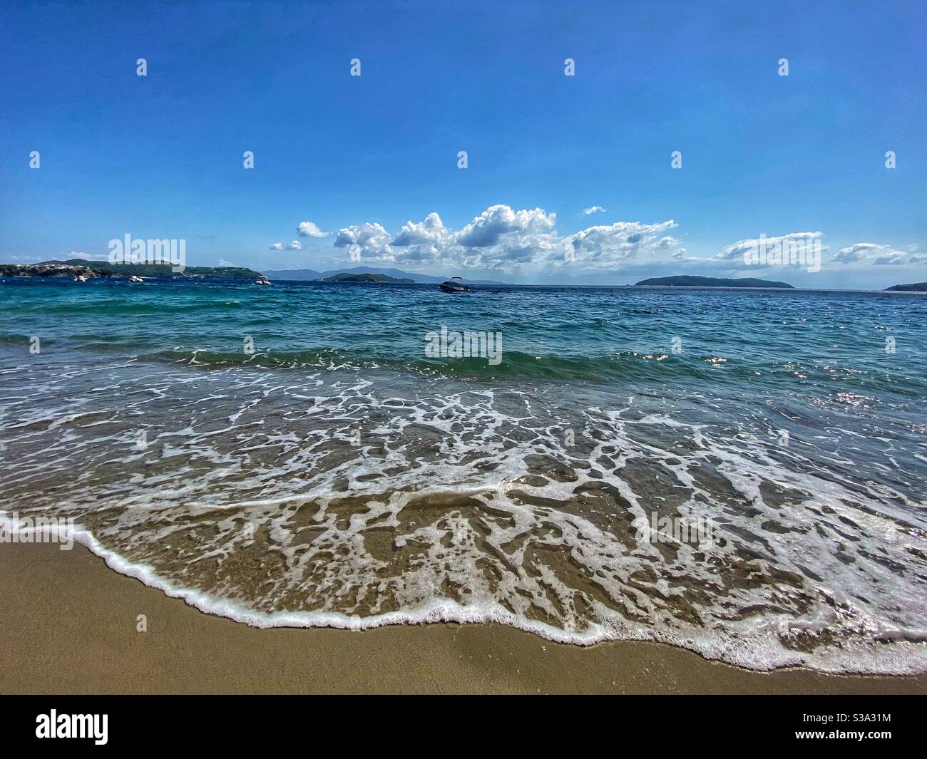 Mare con schiuma sulla sabbia, barche e piccole isole verdi sullo sfondo dell'isola di Skiathos in Grecia. Foto Stock