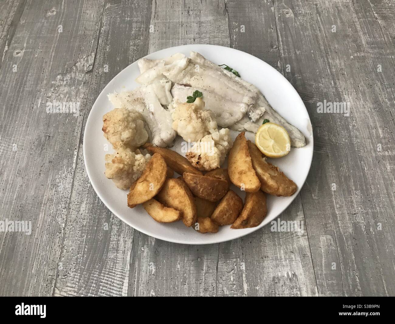 Un piatto di eglefino alla griglia, zeppe di patate e cavolfiore arrosto su sfondo di legno - Immagine stock catturata con smartphone