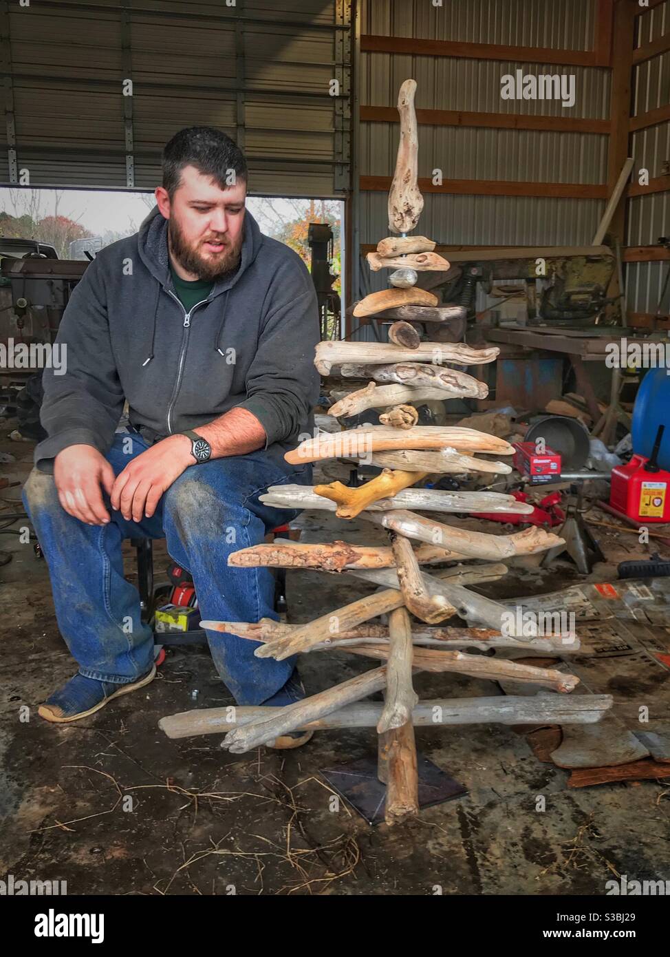 Il giovane uomo bearded ha posato accanto l'albero di Natale di driftwood appena assemblato Foto Stock