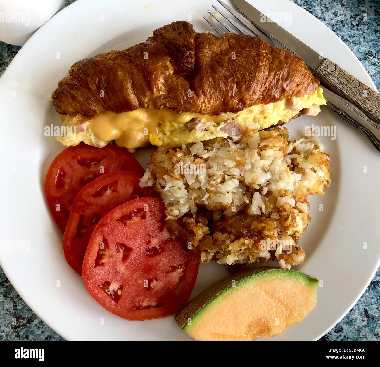 Croissant ripieno con uova e omelette al formaggio, patate fritte, pomodori a fette e melone su un piatto. - Immagine stock catturata con smartphone