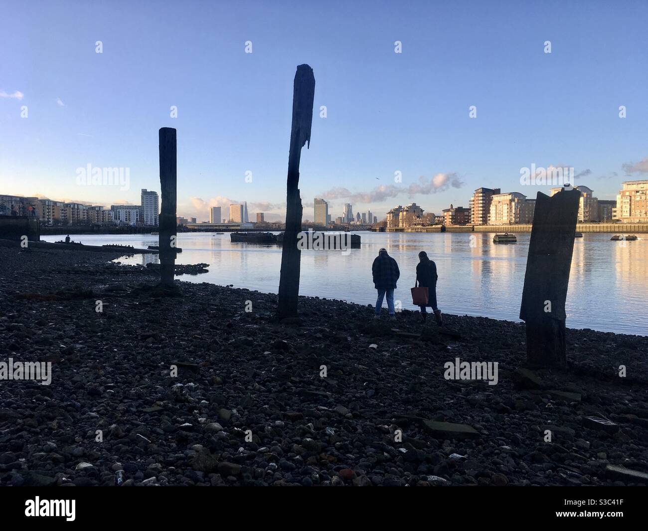 Il Tamigi scorre verso il basso mentre il sole tramonta su londra. 2 persone camminano lungo la riva di Greenwich. Attraverso il fiume Canary Wharf si può vedere dietro edifici privati che si riflettono in acqua. Foto Stock