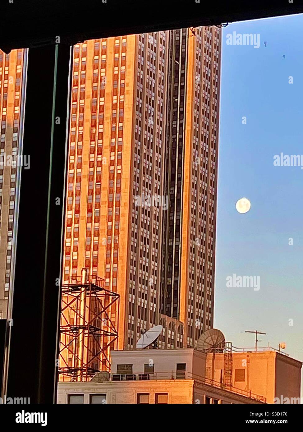 Il Wolf Moon si innamora intorno all'Empire state Building, mentre gli edifici circostanti si illuminano calorosamente alla luce del sole della mattina presto, Manhattan, New York City, USA, visto da una finestra di appartamenti nelle vicinanze. Foto Stock