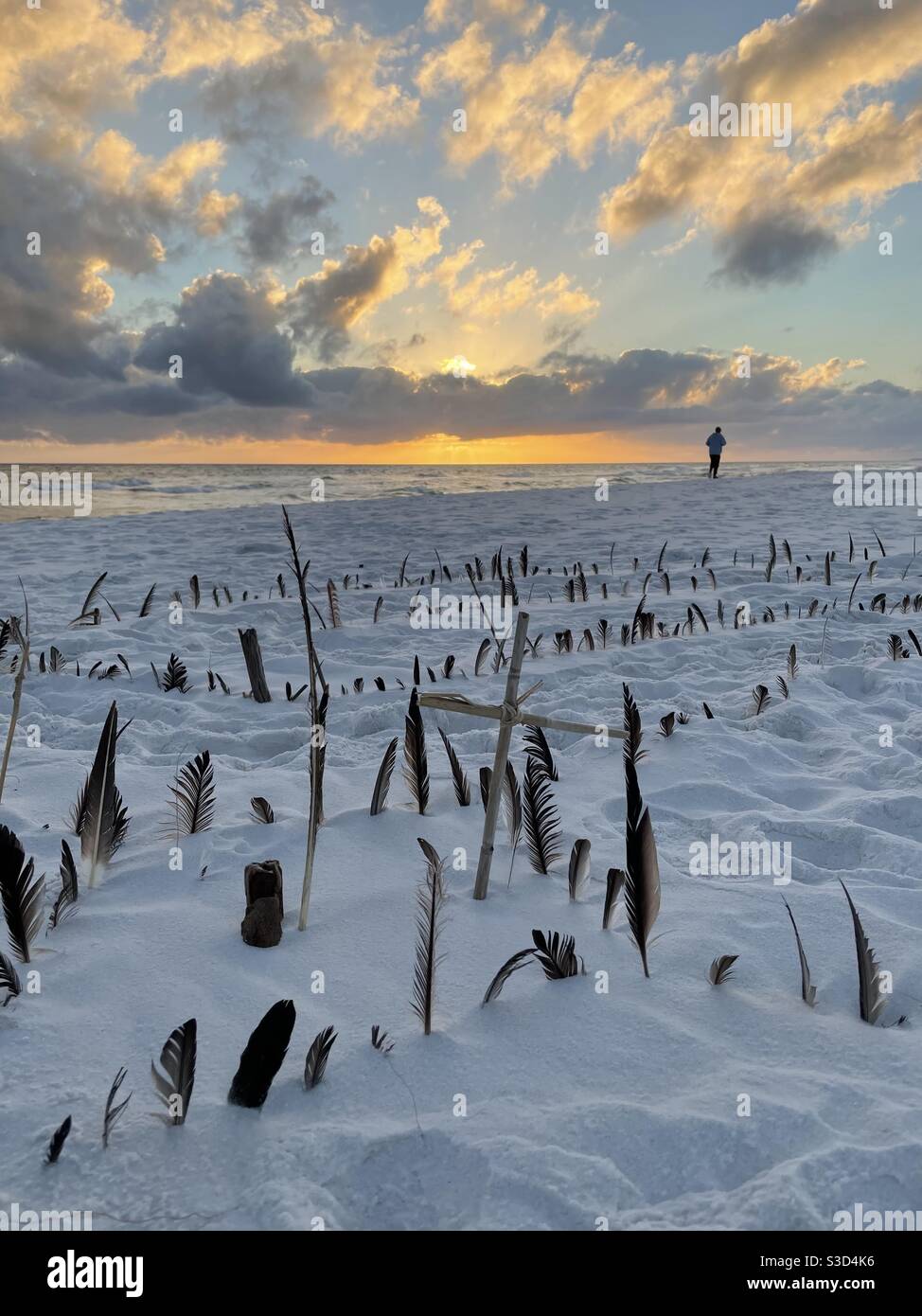 Tramonto sulla spiaggia con un labirinto di piume e croce e silhouette di persona che cammina sulla spiaggia Foto Stock