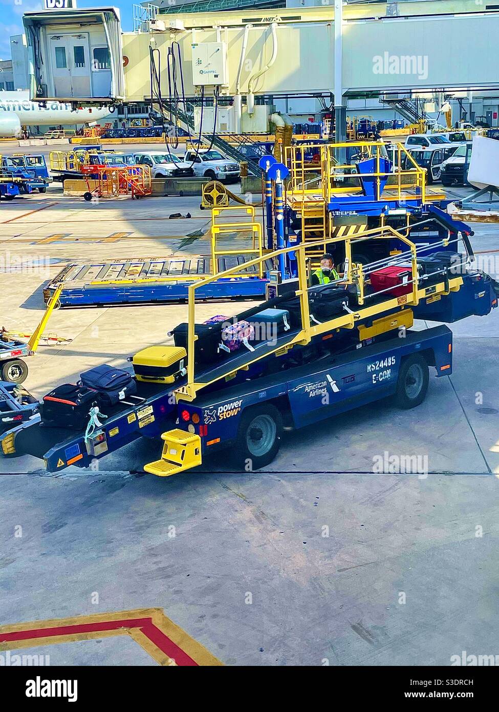 Un lavoratore carica il portaravalige giallo brillante e blu pieno di bagagli colorati passeggeri sulla rampa jetway al terminal American Airlines nell'aeroporto internazionale di Miami, Florida, USA Foto Stock