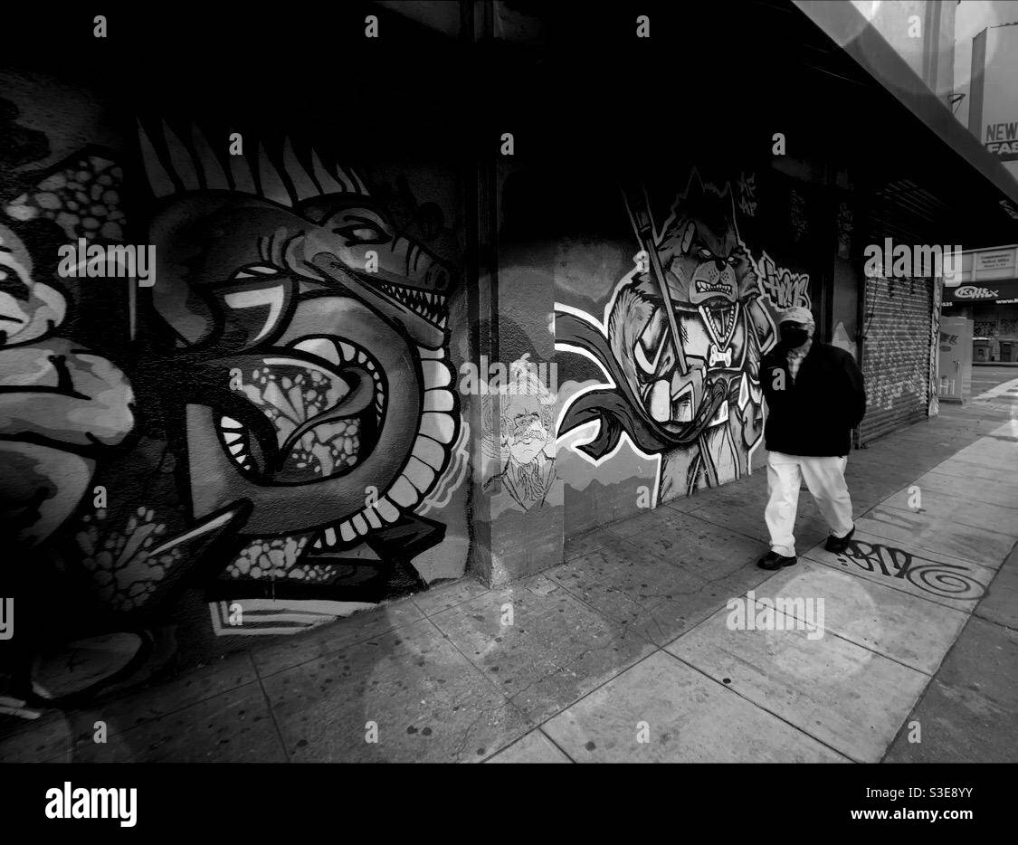 Un uomo che indossa la maschera facciale sta camminando attraverso il marciapiede con i murali del cane arrabbiato e del male serpente al suo fianco Foto Stock