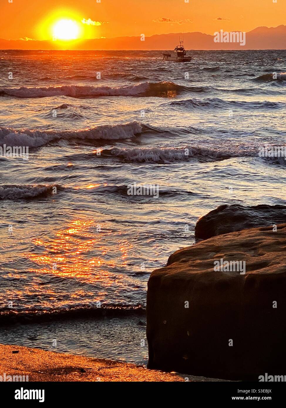 La barca si imbarca su un mare agitate al tramonto. Contrasto del cielo arancione con onde scintillanti metallizzate. Foto Stock