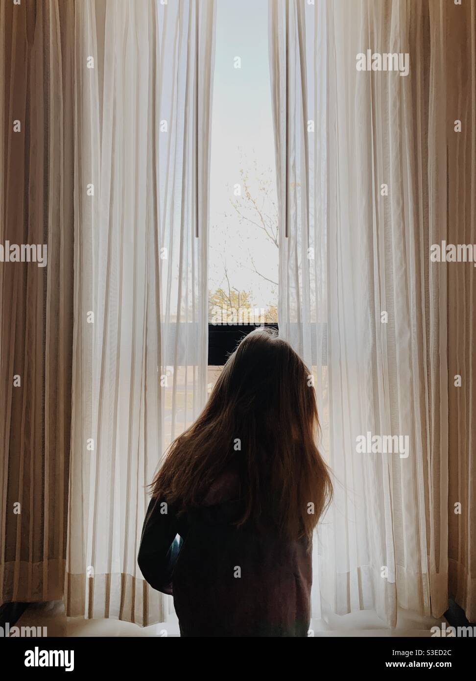 Un bambino piccolo che guarda fuori da una finestra alta con tende bianche in un hotel. Foto Stock