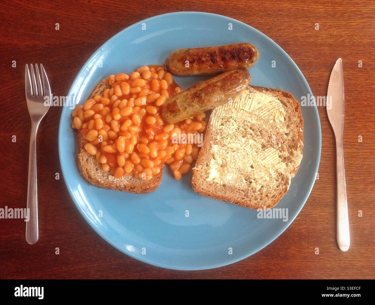 Una colazione cucinata con fagioli su toast, una fetta di pane marrone e salsicce vegetariane su un piatto blu Foto Stock