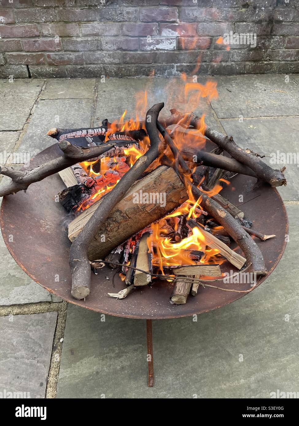Legno che brucia in buca di fuoco arrugginito su pietre del patio vicino muro di mattoni - Immagine stock catturata con smartphone