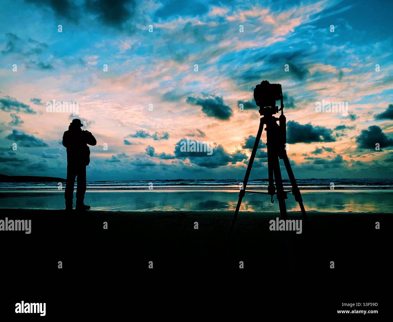 Un uomo scatta foto con una fotocamera su un treppiede Al tramonto su una spiaggia in Cornovaglia - Immagine stock catturata con smartphone