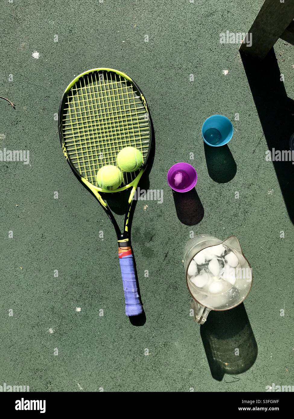 Vista a volo d'uccello delle racchette da tennis su un campo da tennis con palle da tennis a fianco una caraffa di ghiaccio freddo acqua e un paio di tazze su un caldo sole giorno Foto Stock