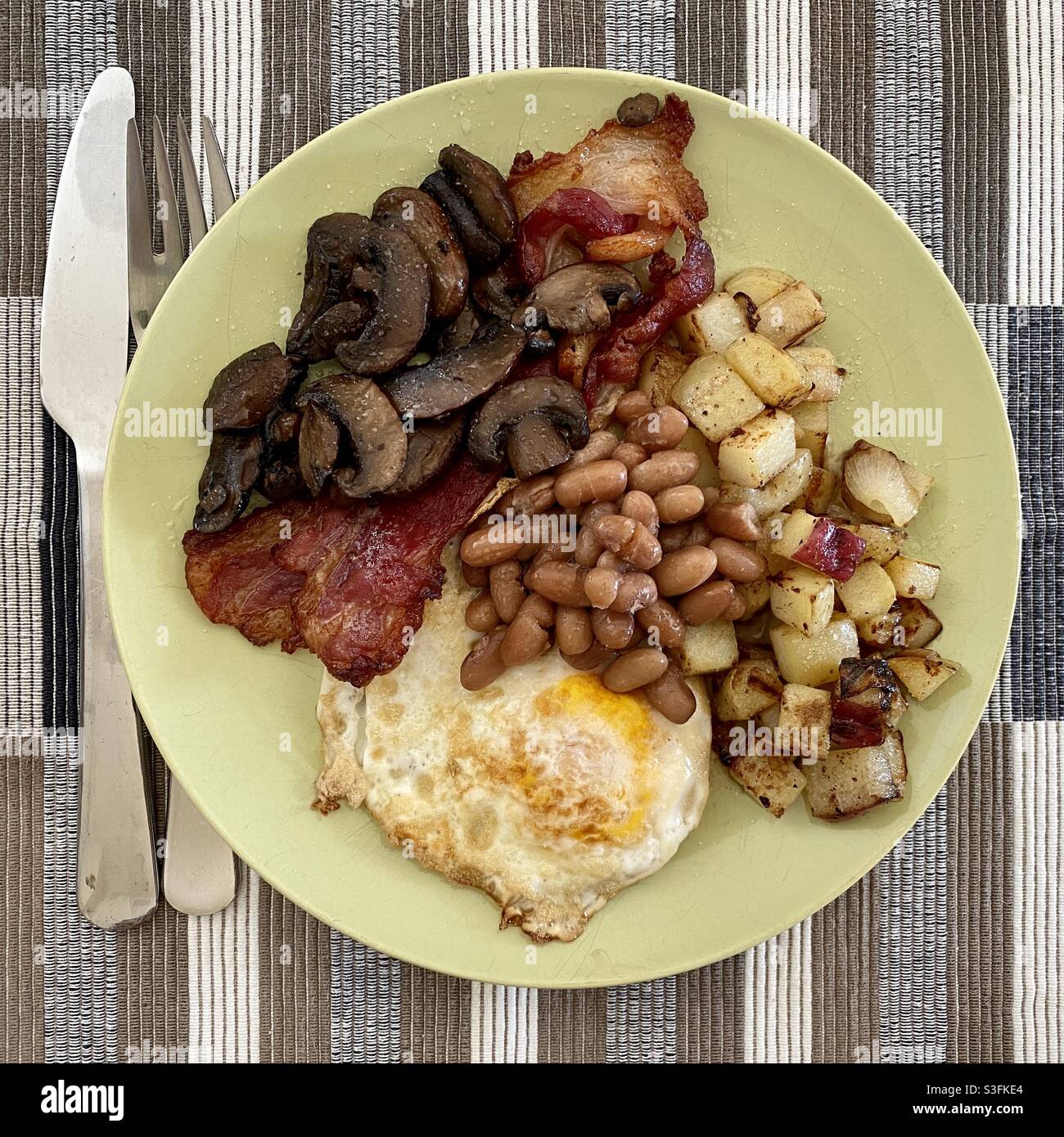 Colazione fritta con uova, pancetta, patate fritte, funghi e fagioli cotti su un piatto verde con posate in acciaio inossidabile a lato Foto Stock