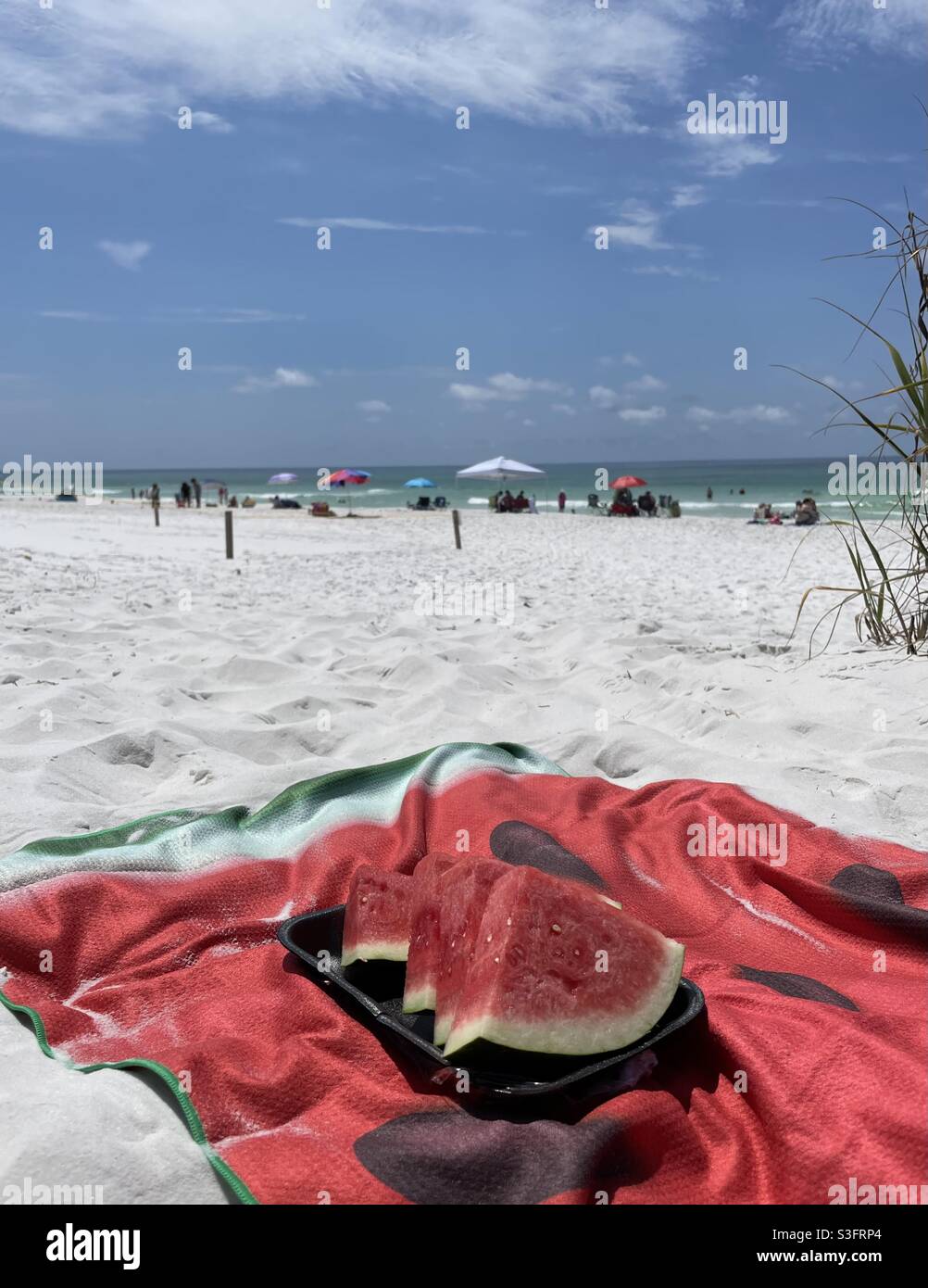 Selezionate la messa a fuoco sul cocomero affettato su un telo da spiaggia rosso con persone sfocate sulla spiaggia di sabbia bianca Foto Stock