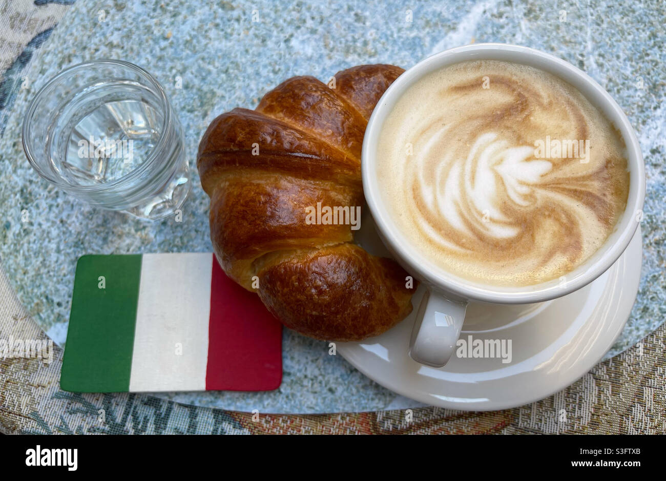 Classica colazione italiana al bar con cappuccino, croissant, un bicchiere d'acqua e la bandiera italiana Foto Stock
