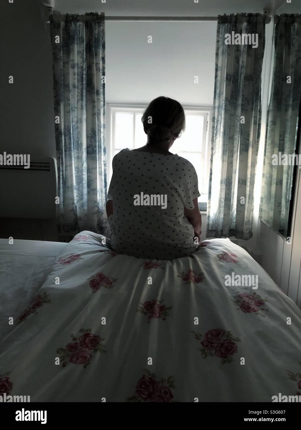 Un uomo donna solitario seduto da solo nella sua camera da letto e guardando fuori da una finestra in un concetto di depressione, ansia o solitudine - Immagine stock catturata con smartphone