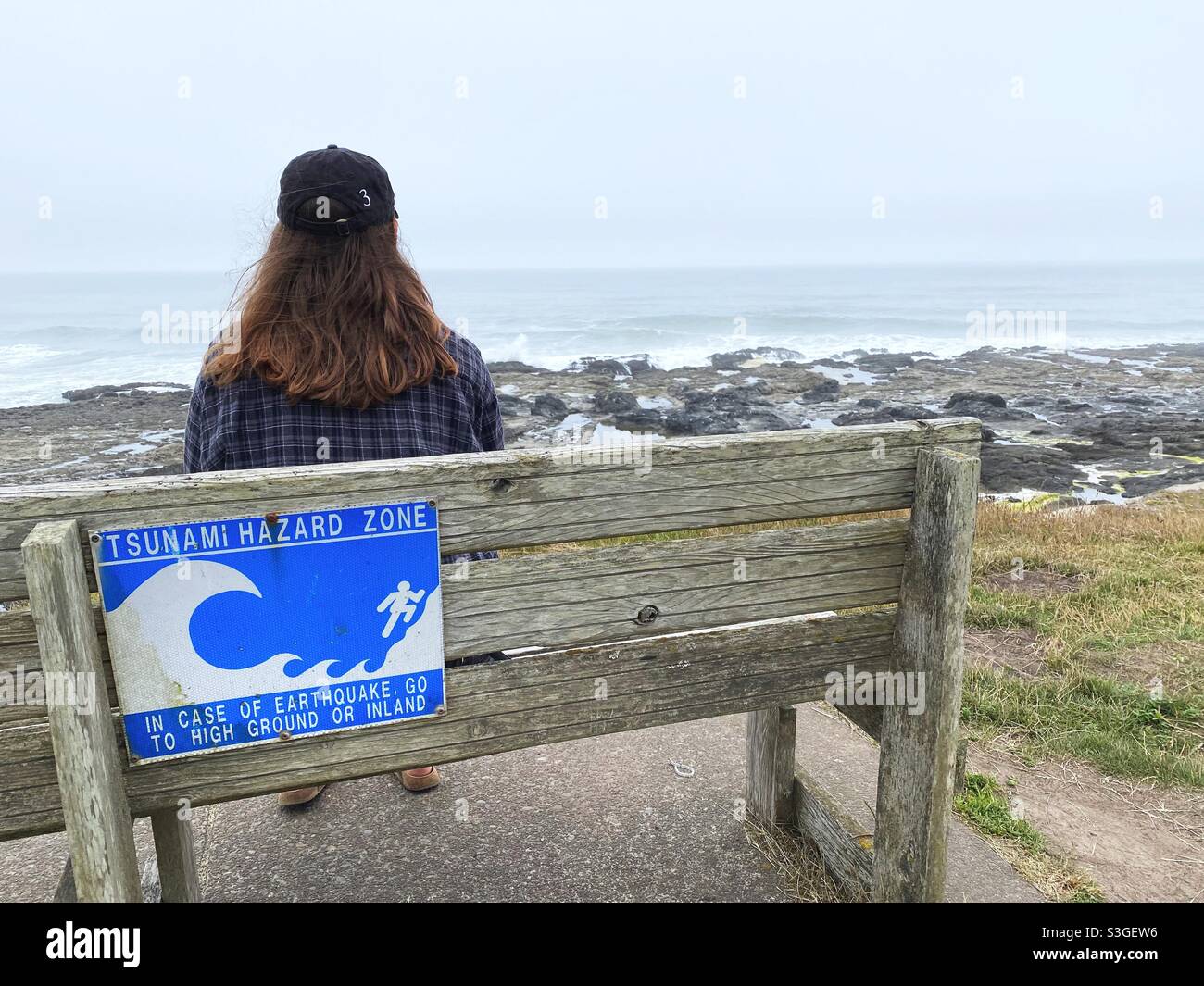 Una persona seduta su una panchina all'oceano con un cartello di avvertimento dello tsunami, a Yachats, Oregon. Foto Stock