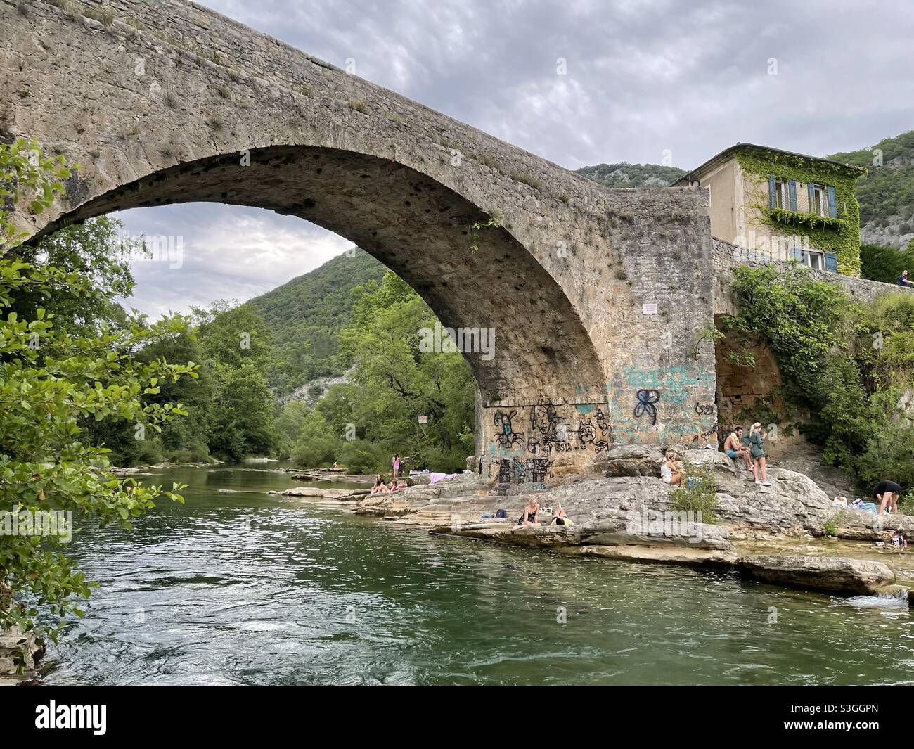 I bagnanti si siedono sulla riva sotto un grande ponte di pietra con una vecchia casa di villaggio sul fiume Hérault vicino a Ganges e Montpellier, Occitanie, a sud della Francia - Immagine stock catturata con smartphone