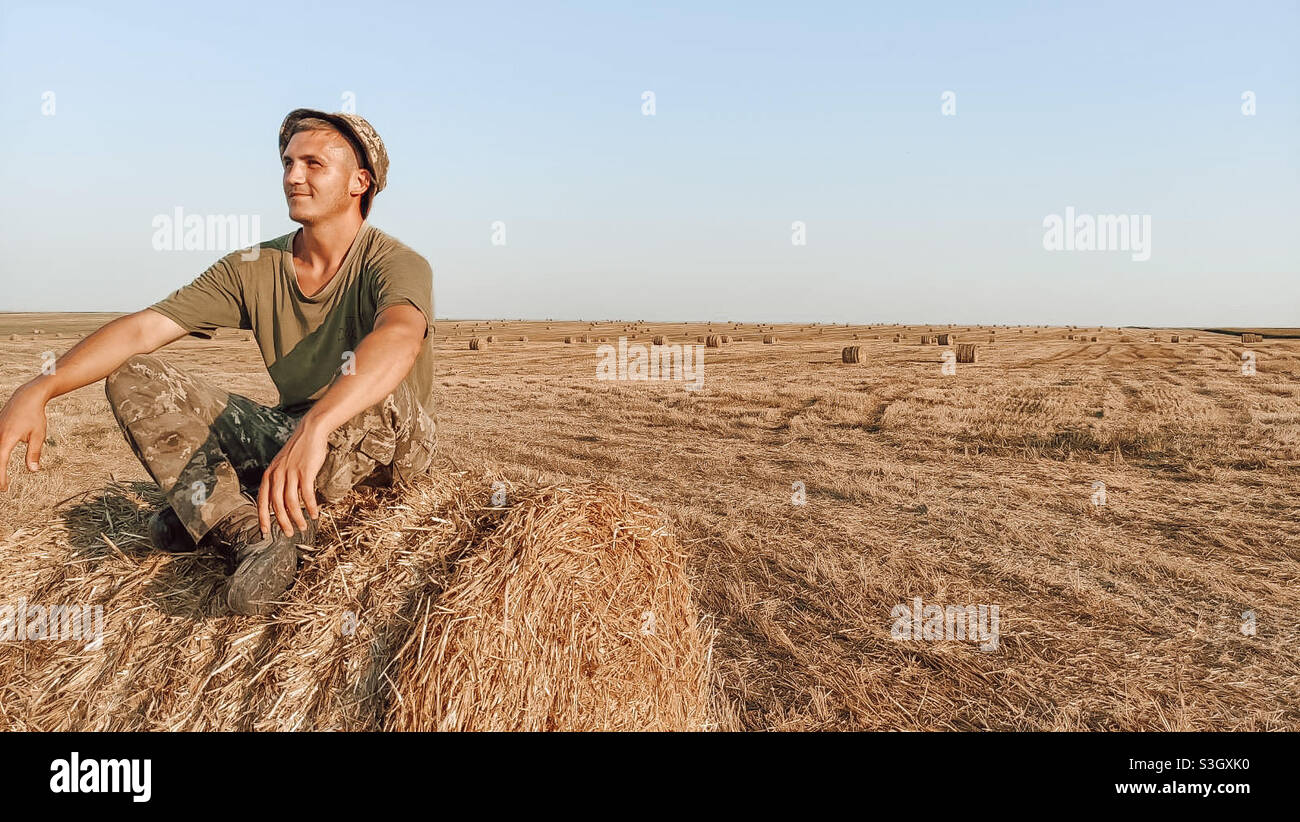 Giovane uomo seduto su un pagliaio nel mezzo di un campo giallo Foto Stock
