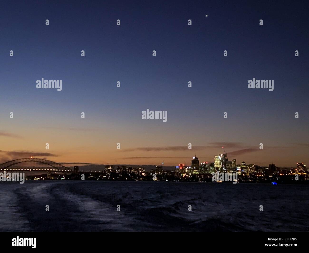Il ponte del porto di Sydney e lo skyline della città di Sydney al tramonto con la stella serale venus che splende in un cielo post-tramonto sulle onde del risveglio di una barca Foto Stock