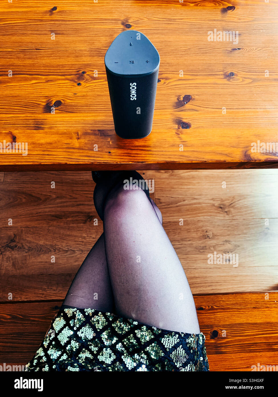 Diffusore portatile Sonos Roam su un tavolo in legno e una donna con gambe incrociate che indossa un abito a paillettes Foto Stock