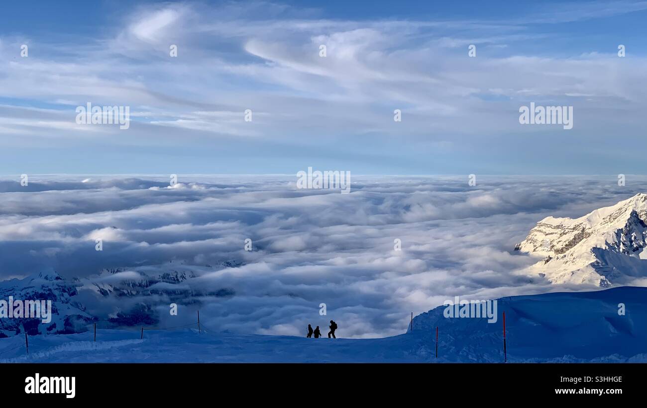 In cima al Monte Titlis guardando il panorama con tre sciatori in procinto di scendere. Engelberg è sotto le nuvole. Foto Stock