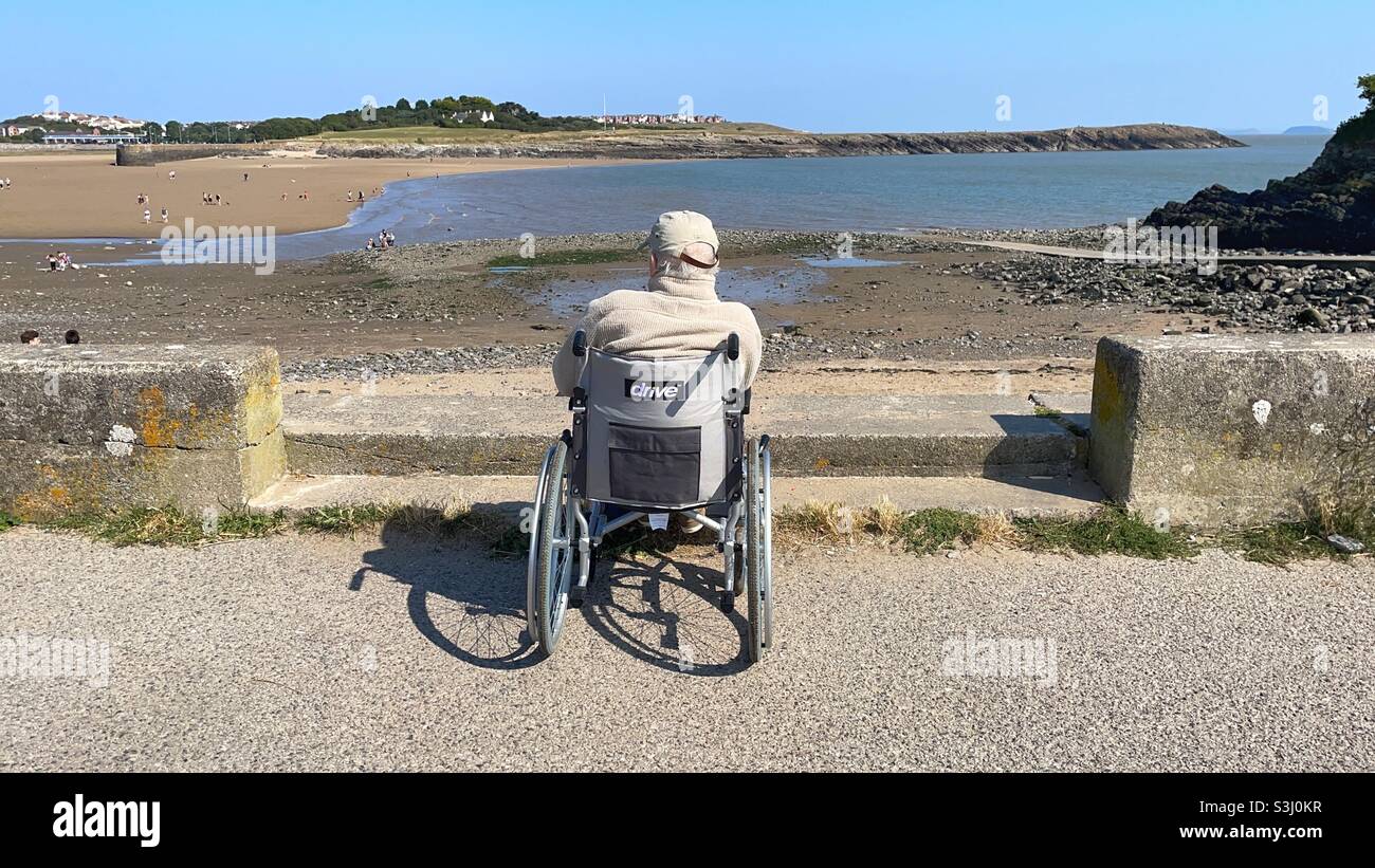 Uomo anziano in sedia a rotelle che guarda il mare in una giornata di sole. Foto Stock