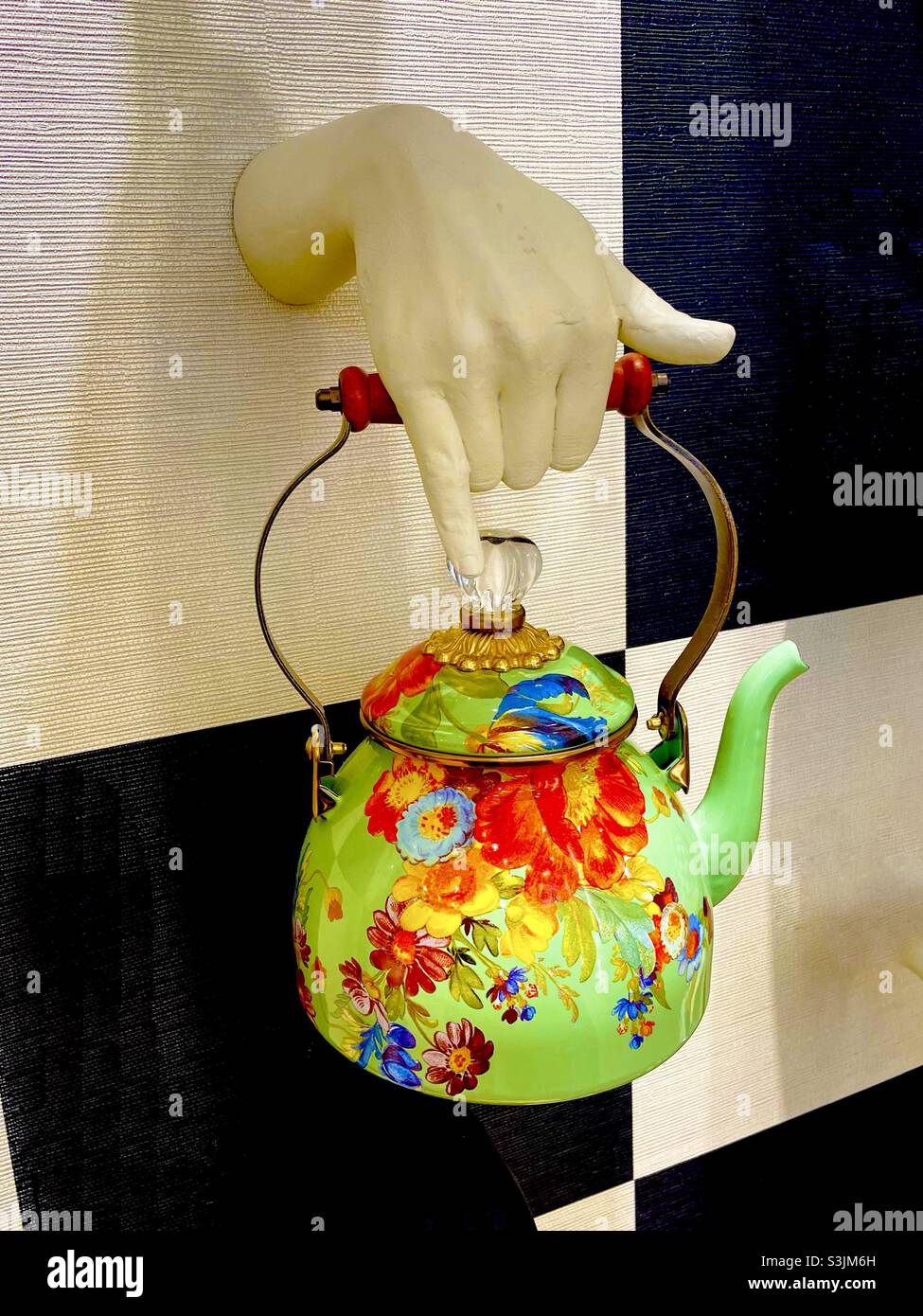 Iconica pentola da tè verde floreale Mackenzie Childs su una parete in piastrelle bianche e nere che viene tenuta da una mano in ceramica bianca - Immagine stock catturata con smartphone