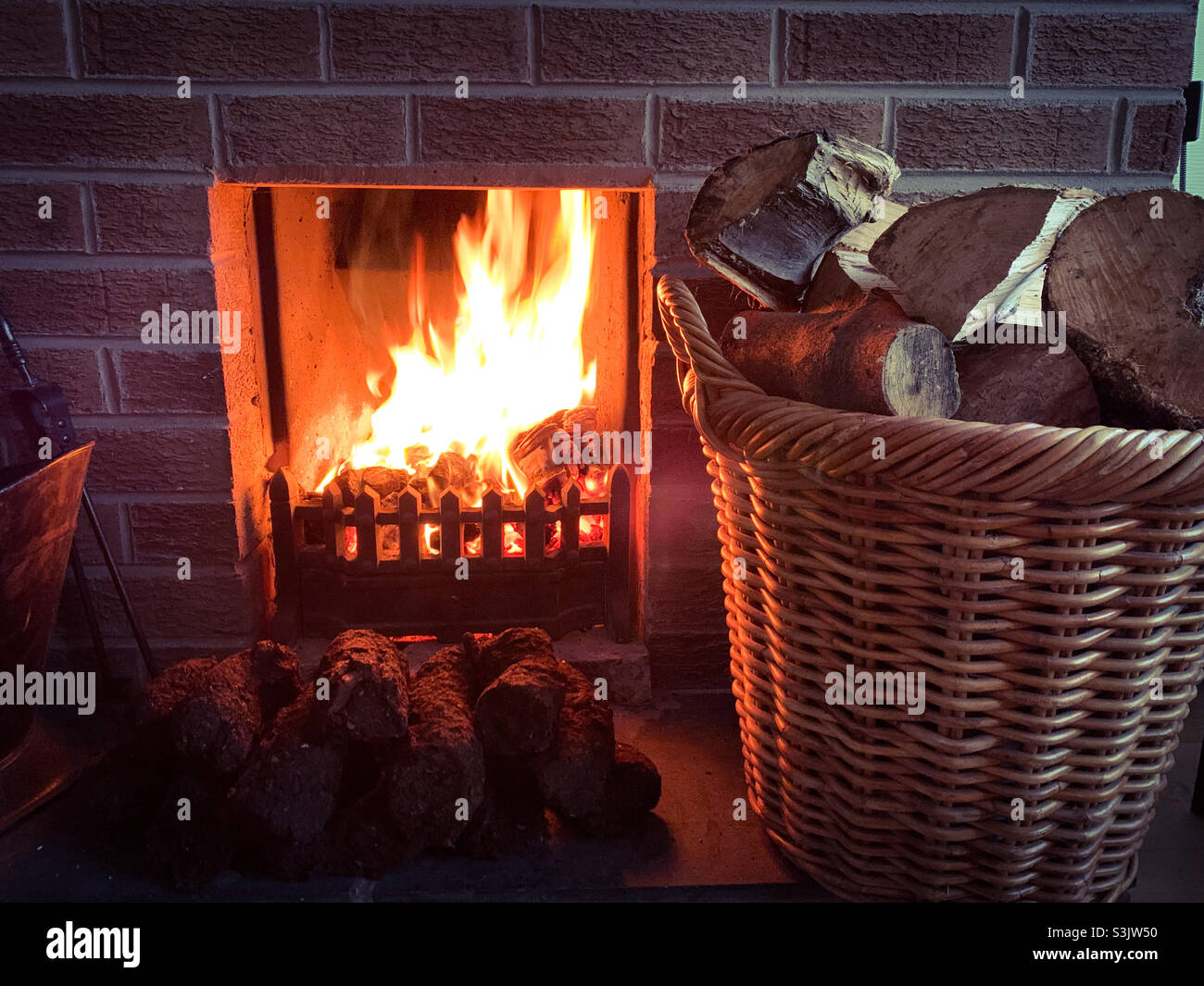 Un fuoco di erba sintetica ben illuminato con tronchi in un cesto e tappeto erboso che asciuga sul focolare. Foto Stock