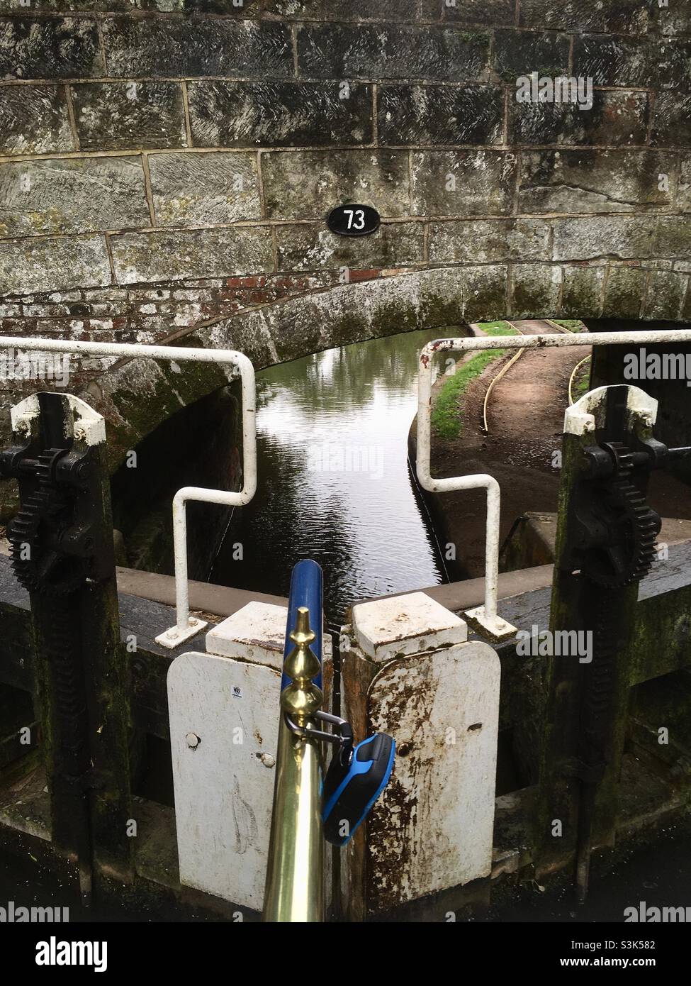 Ponte del canale con targhetta di identificazione vista dalla poppa di un battello narrowboat che sale una serratura con i cancelli di bloccaggio e il perno del timone in primo piano. - Immagine stock catturata con smartphone