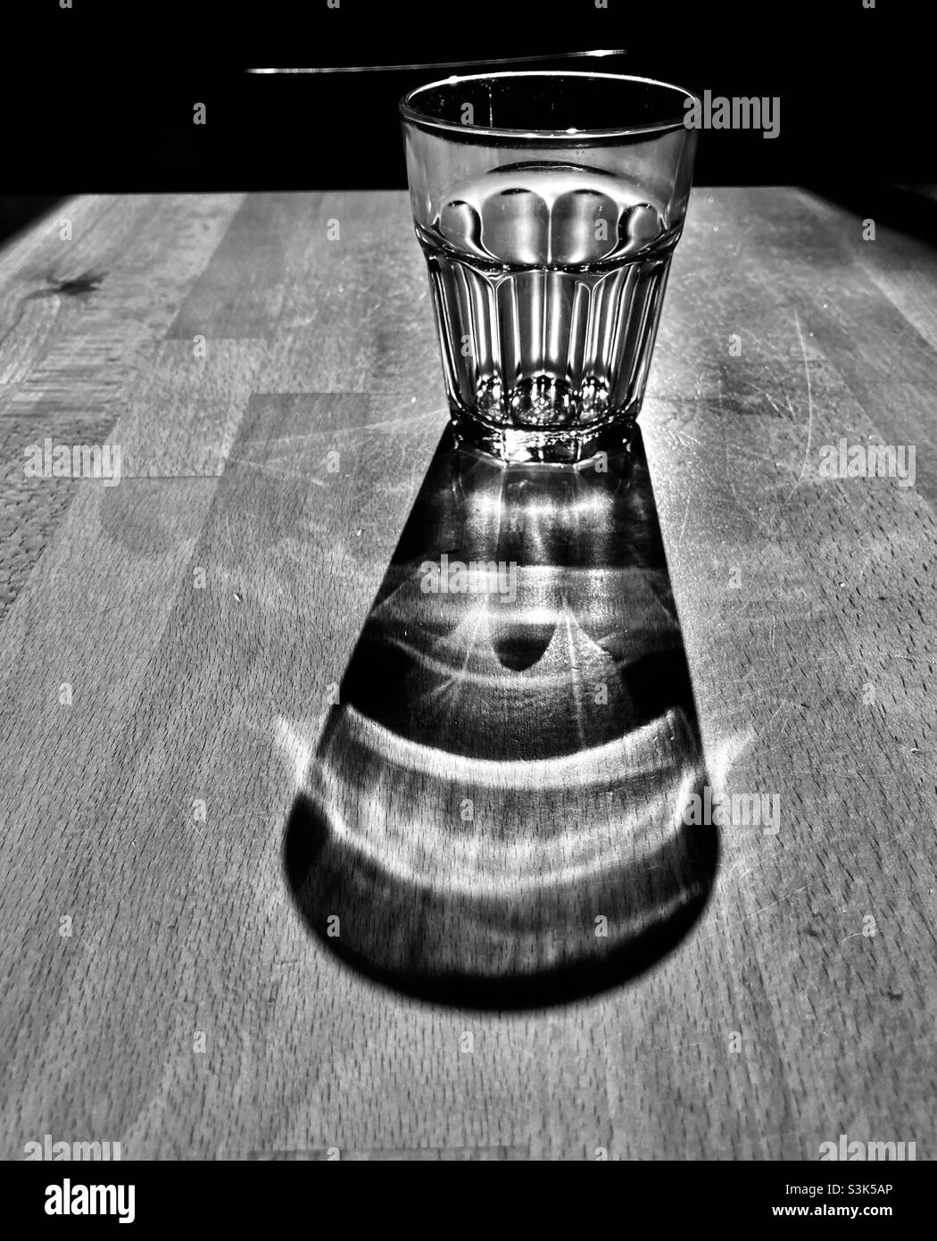 un bicchiere d'acqua si erge su un tavolo di legno e getta una lunga ombra Foto Stock