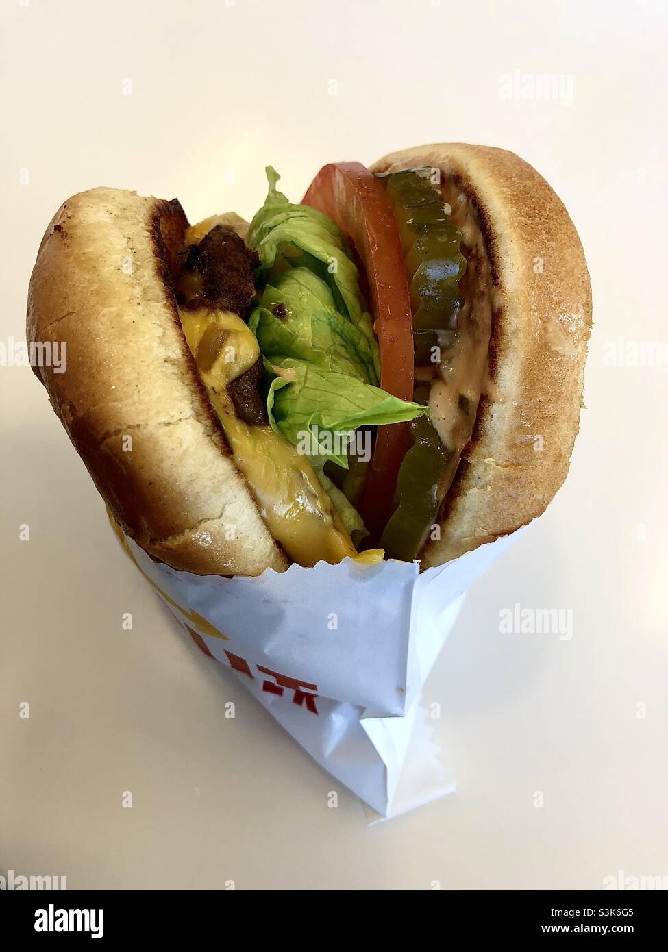 Un cheeseburger in confezione bianca con fondo neutro e vuoto. Alcuni colori rosso e giallo non leggibili vengono stampati sulla pellicola. Sottaceti, pomodori, lattuga, cipolle cotte all'interno della panna sono tutti molto visibili. Foto Stock