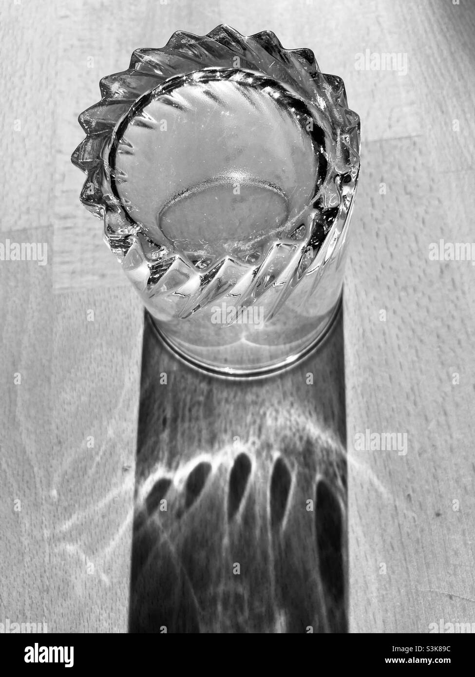 un bicchiere d'acqua si erge su un tavolo di legno e getta una lunga ombra Foto Stock