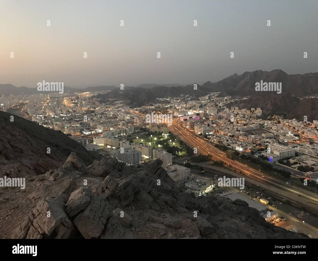 Crepuscolo sopra una città: Una vista dalla cima di una collina. Oman, Greater Muttrah e wadi Kabir illuminati con luci serali. - Immagine stock catturata con smartphone