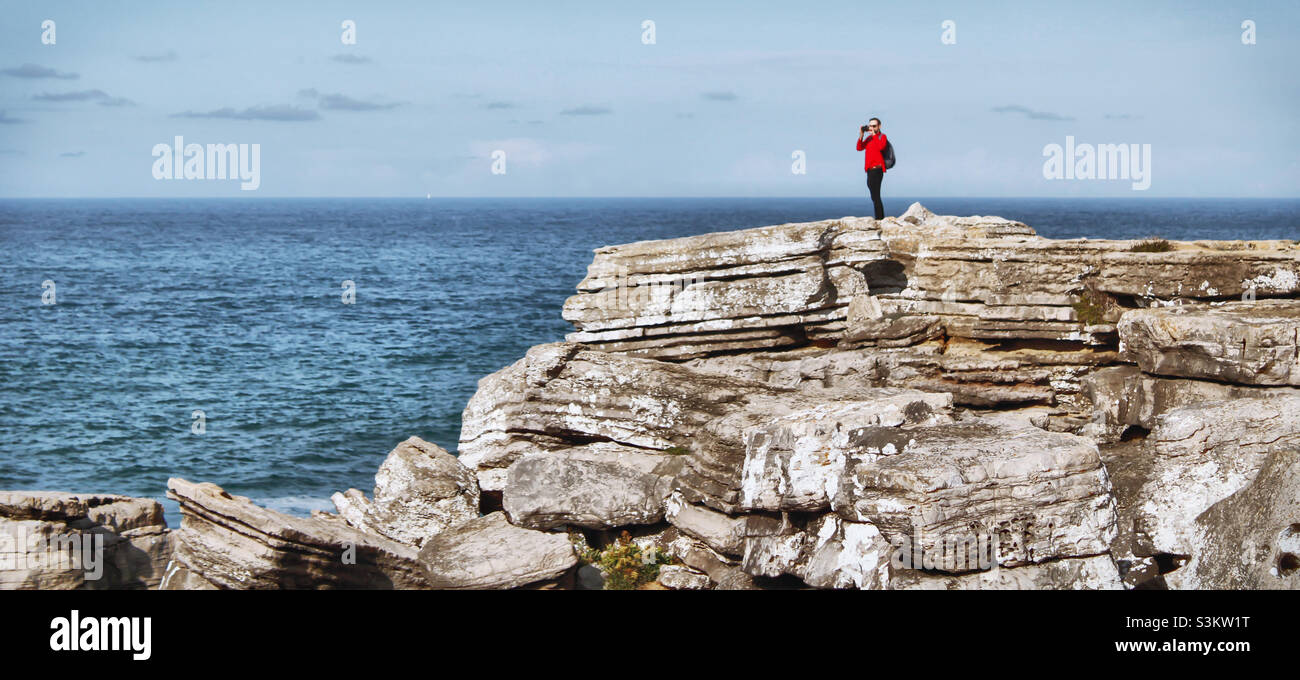 Un uomo in una camicia rossa si erge su un affioramento roccioso scattando foto accanto all'Oceano Atlantico a Peniche, Portogallo Foto Stock