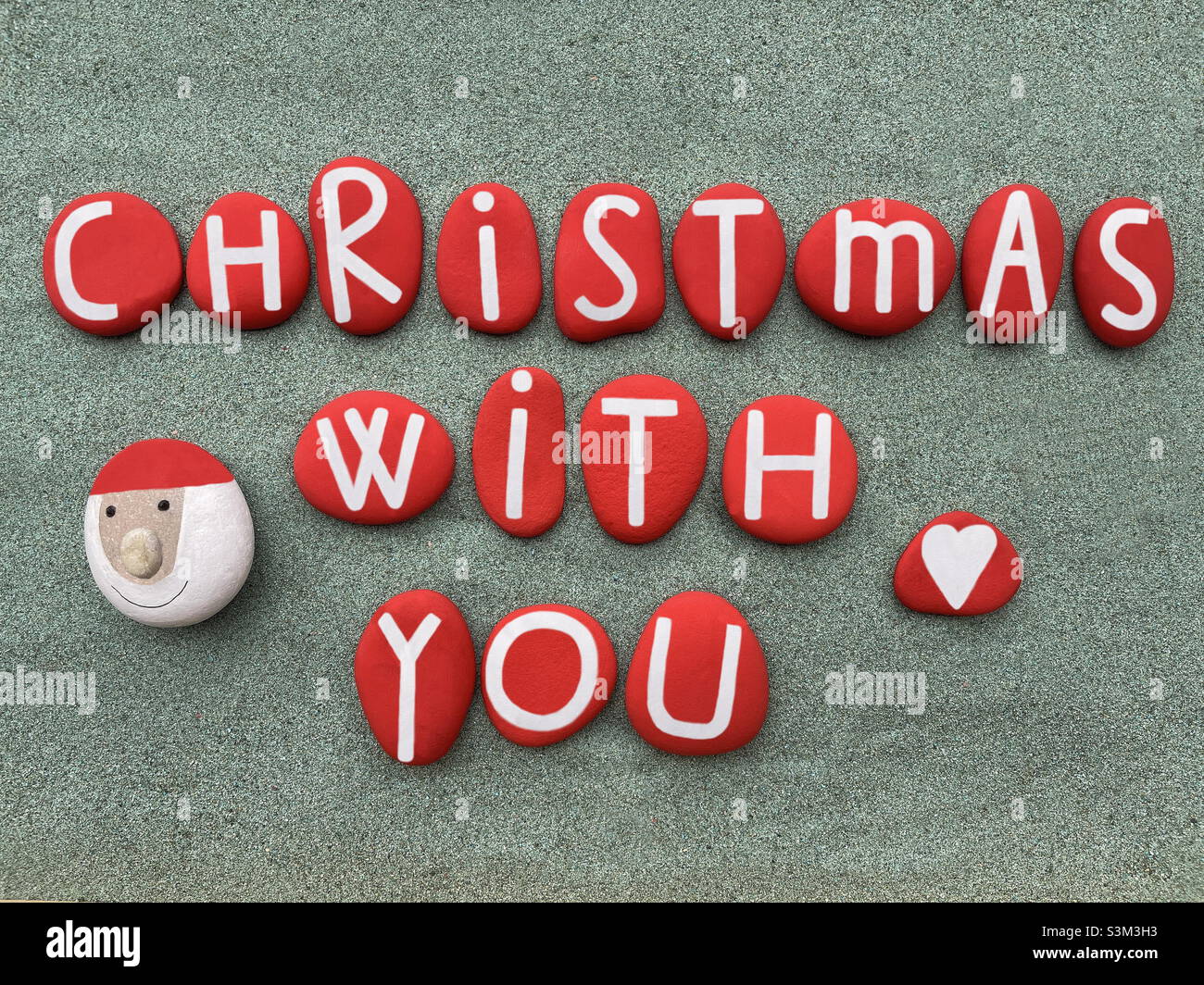 Natale con voi, frase creativa composta con lettere di pietra rossa dipinte a mano e s pietra Babbo Natale su sabbia verde - Immagine stock catturata con smartphone