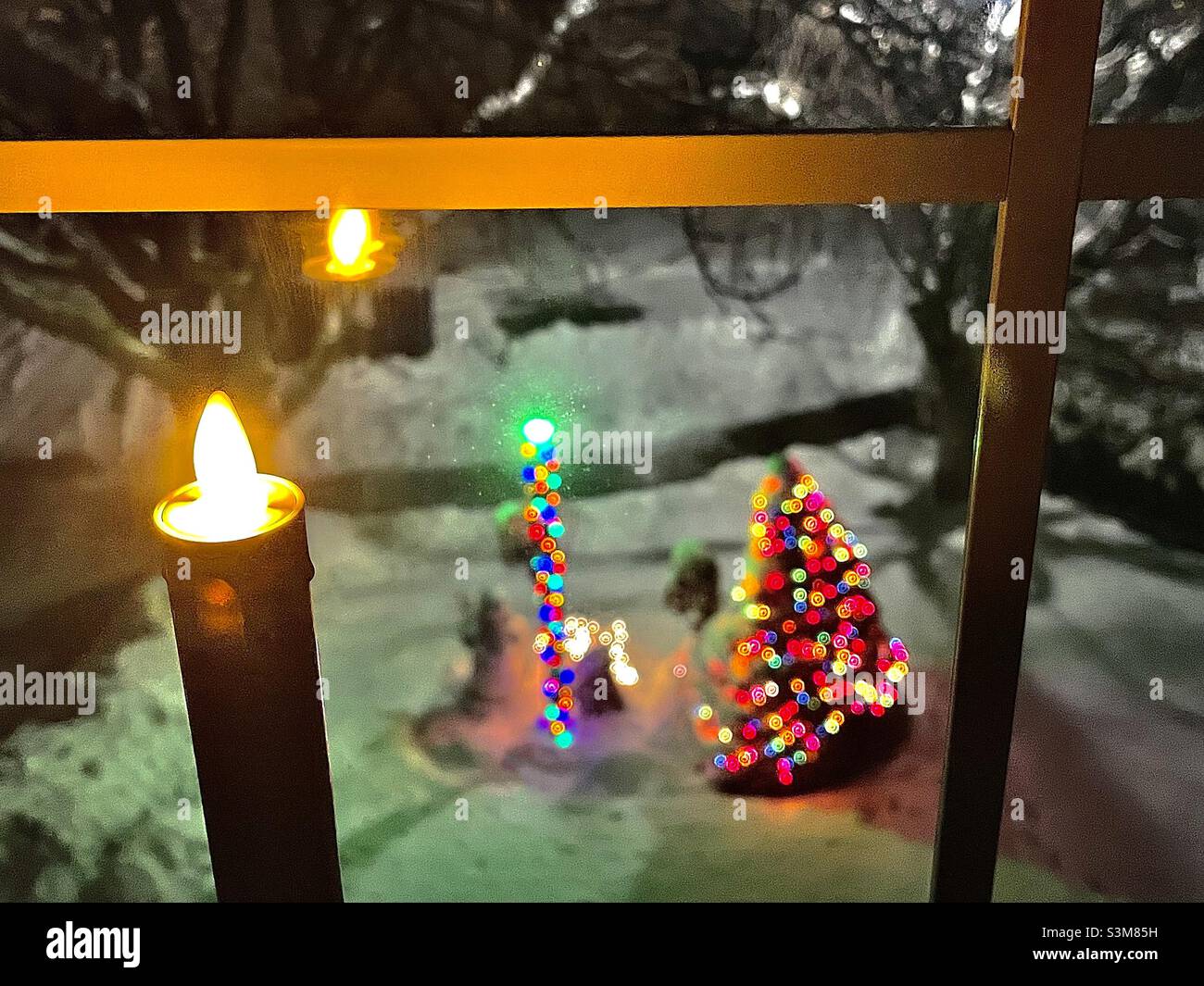 Una candela a batteria si illumina e sfarfalla nella finestra di una casa al piano superiore, guardando fuori nel loro cortile con le luci di Natale e la nuova nevicata in Utah, Stati Uniti. - Immagine stock catturata con smartphone