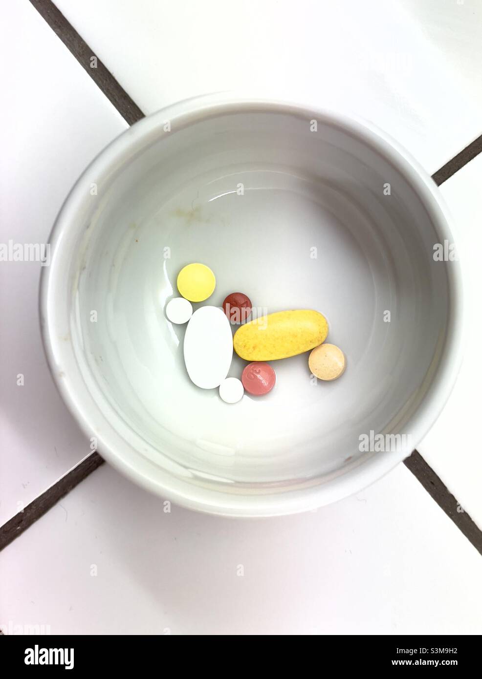Un assortimento di pillole e compresse in un piccolo recipiente in ceramica bianca su un piano di lavoro piastrellato Foto Stock