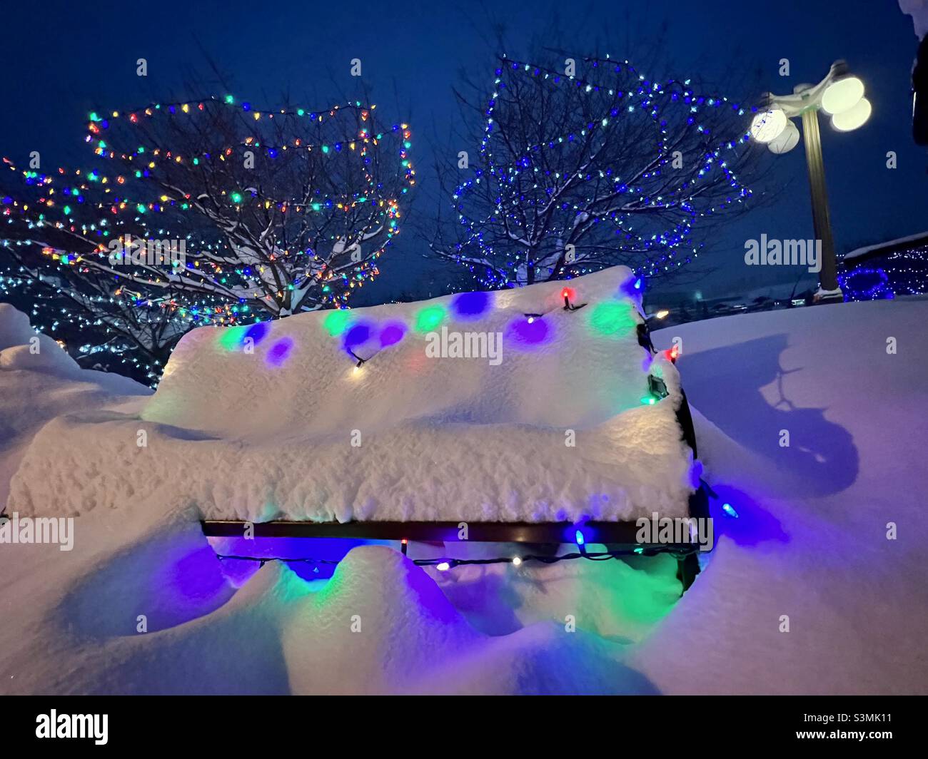 Panca di parco coperta di neve profonda con luci colorate di Natale di notte Foto Stock
