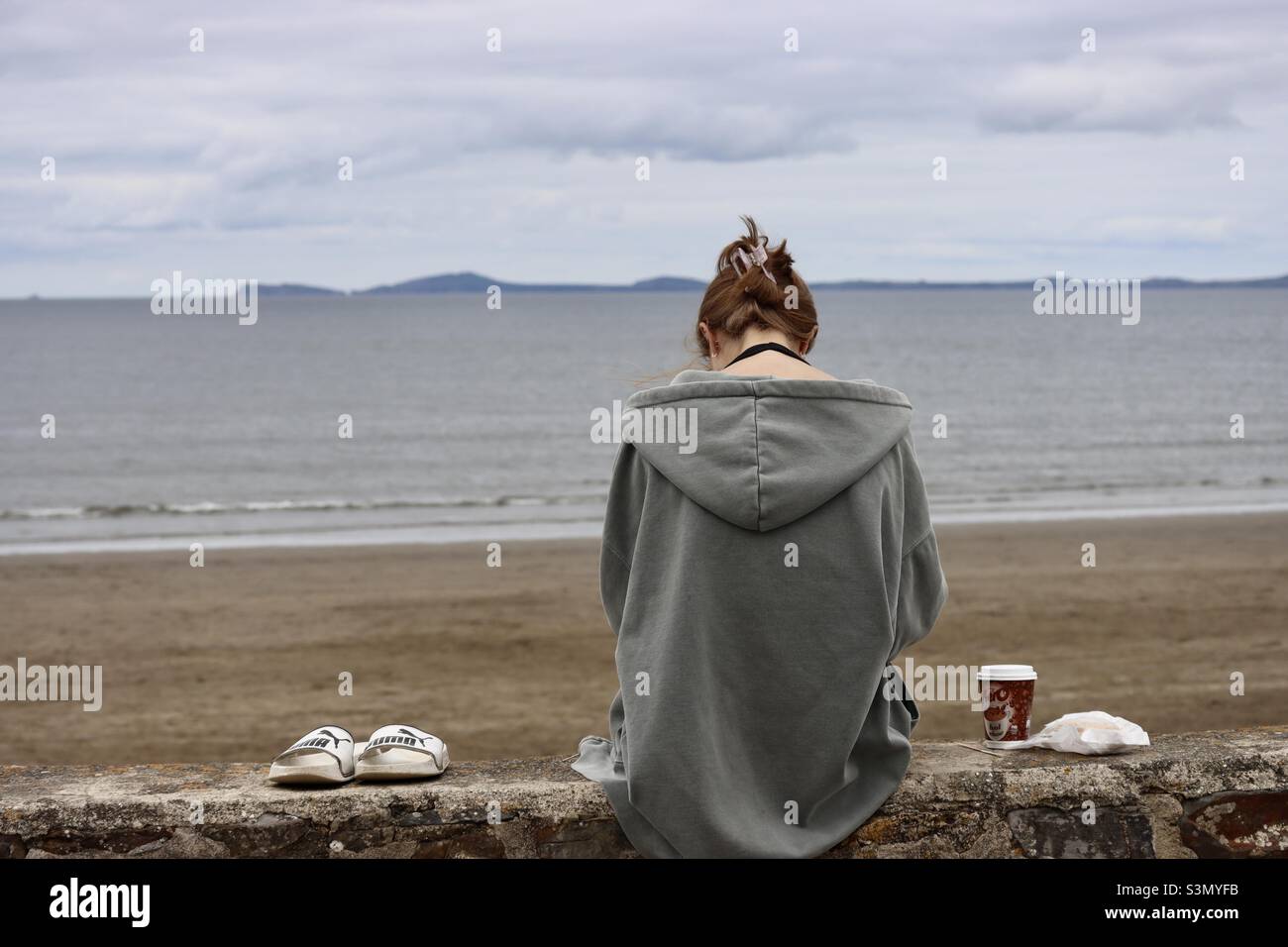 Una signora da dietro, seduta su un muro in spiaggia. Godendo un caffè con le sue scarpe sul muro oltre a lei. Lei sta portando i suoi capelli legati e ha un oversize felpa con cappuccio su. Foto Stock