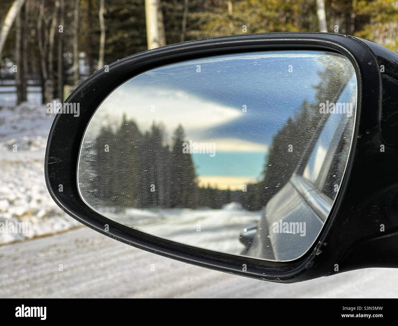 Una lunga strada ricoperta di neve e alberata con pesanti nubi ad arco chinook, come si vede nello specchietto retrovisore esterno di un veicolo. Ai piedi dell'Alberta, Canada. Foto Stock