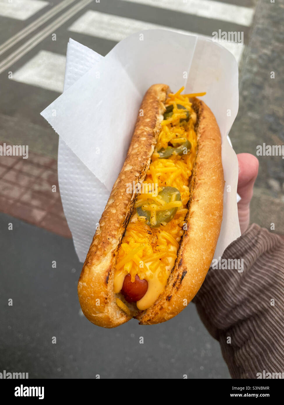 Maschio mano che tiene hot dog con jalapenos e cheddar alla griglia in un tovagliolo di carta all'aperto sulla strada. Foto Stock