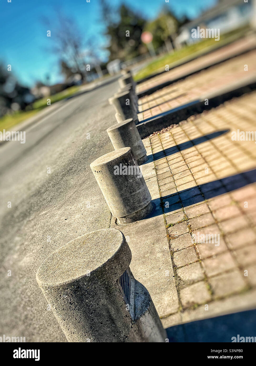 Una vista inclinata in una giornata di sole di una linea di barriere circolari in cemento che separano un passaggio pubblico da una strada, con lunghe e strette ombre che si estendono da ciascuna di esse. Foto Stock