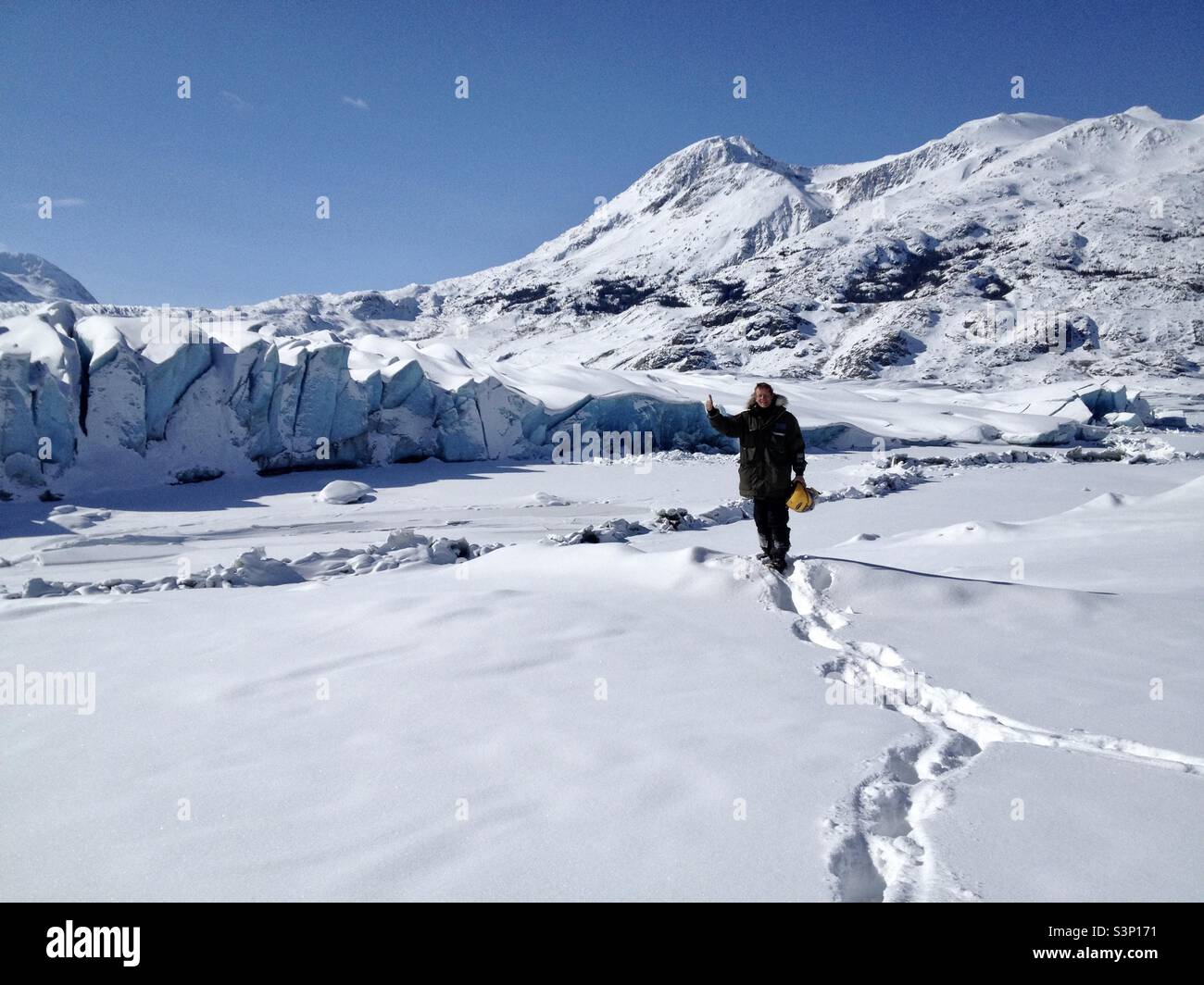 Uomo con parka invernale e casco giallo che dà le dita davanti al ghiacciaio blu con montagne innevate e cielo blu - Immagine stock catturata con smartphone