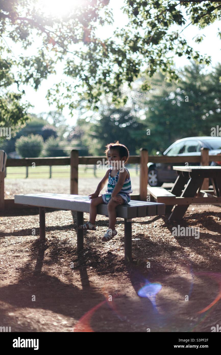 Giovane bambina seduta su una panca del parco in una calda giornata estiva adrenata al sole Foto Stock