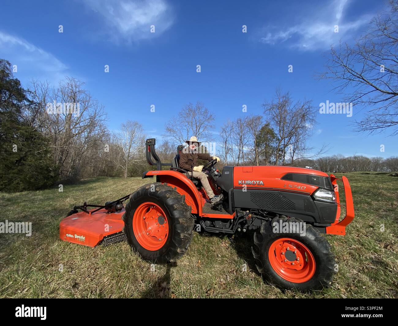Un uomo bianco di mezza età che indossa un cappello a brimmetto largo siede su un grande trattore agricolo arancione nella valle di Shenandoah in Virginia. Foto Stock