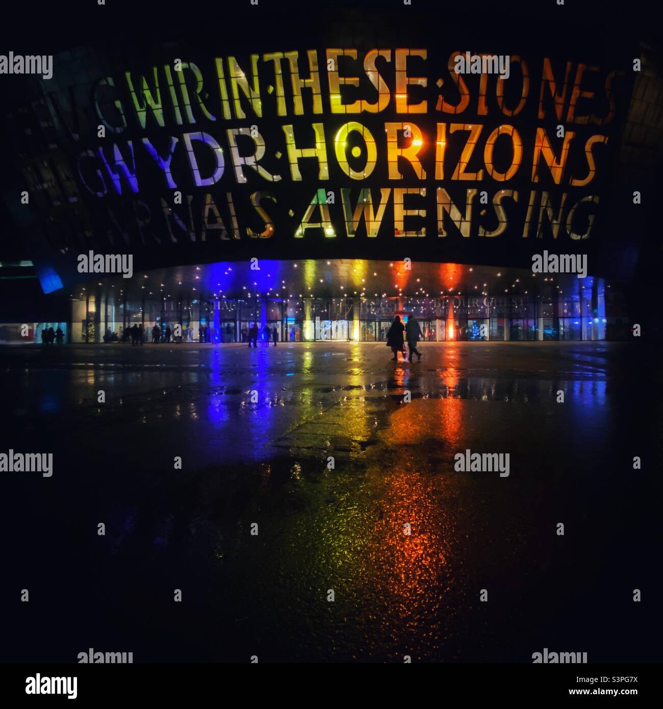 Una fotografia del Wales Millennium Center in una notte di pioggia scura, illuminata in Pride Colors. Riflessioni nelle pozzanghere, e una coppia passeggiando al teatro Foto Stock