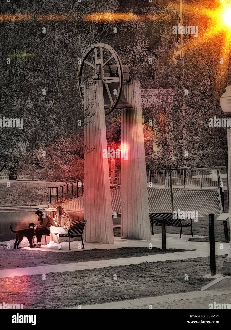 Due amici e il loro cane siedono su una panchina sotto un facsimile Liberty Bell nel Memory Grove Memorial Park a Salt Lake City, Utah, USA. Un'alba aggiunta digitalmente con un tocco di sole aggiunge interesse artistico. Foto Stock