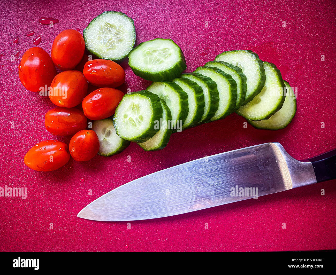 Cetriolo affettato con pomodori pelati con un coltello su un tagliere rosso. - Immagine stock catturata con smartphone